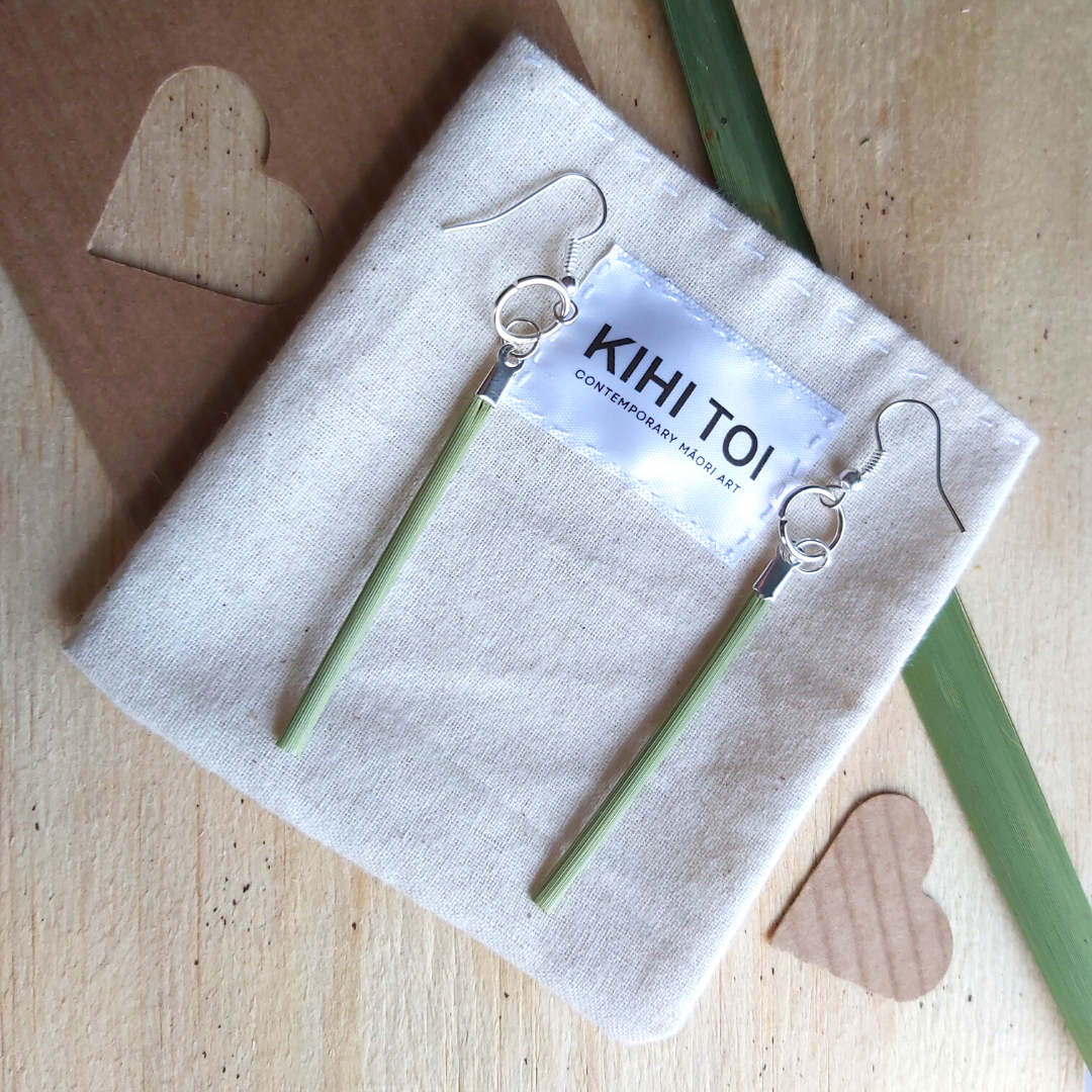 Photo of two earrings placed on top of a KIHI TOI hand sewn calico pouch, each hand fused using a Stainless Steel french hook, a bright silver medium sized jump ring, a small look small sized jump ring, a silver look folded ribbon clamp end folded around a short, thin, dried, green flax leaf piece. A simple, natural, yet eloquent design.